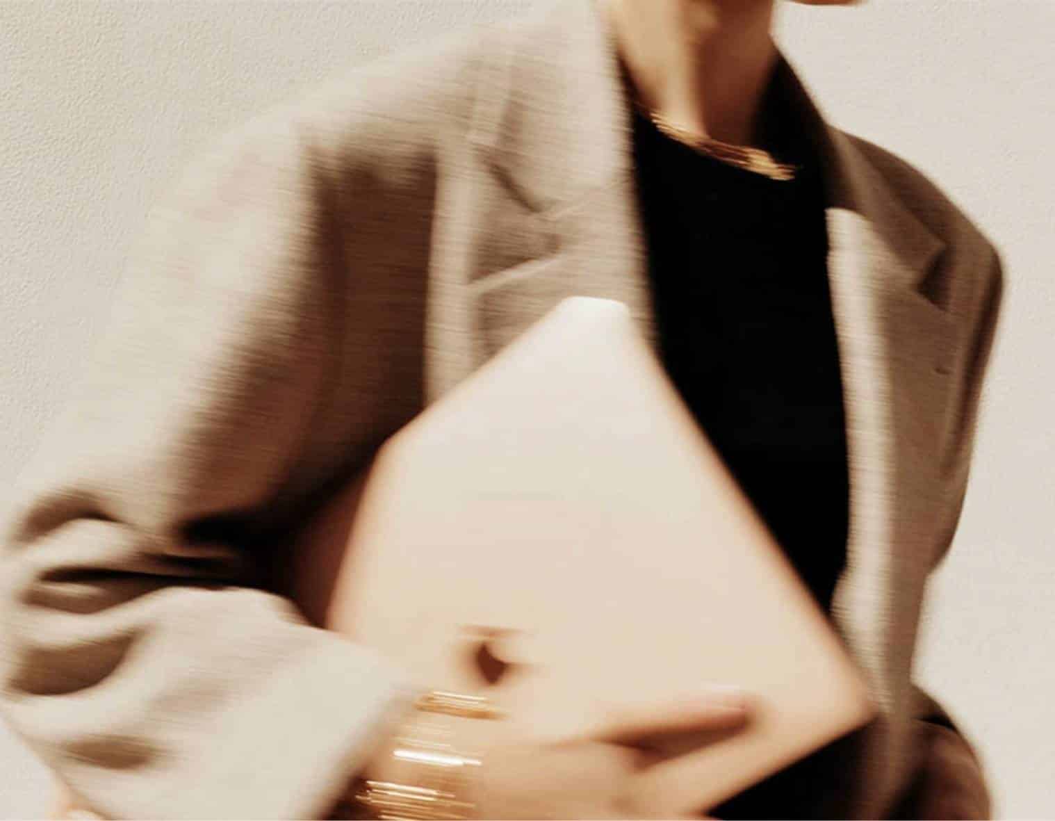 Femme élégante en mouvement tenant un ordinateur portable, blazer beige et bijoux dorés, univers visuel d'un studio de branding beauté et bien-être à Lyon